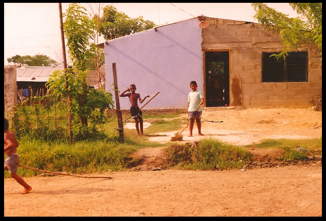 3 Poz&oacute;n boys ride
              rudimentary stick horses in dirt street by cinderblock
              houses