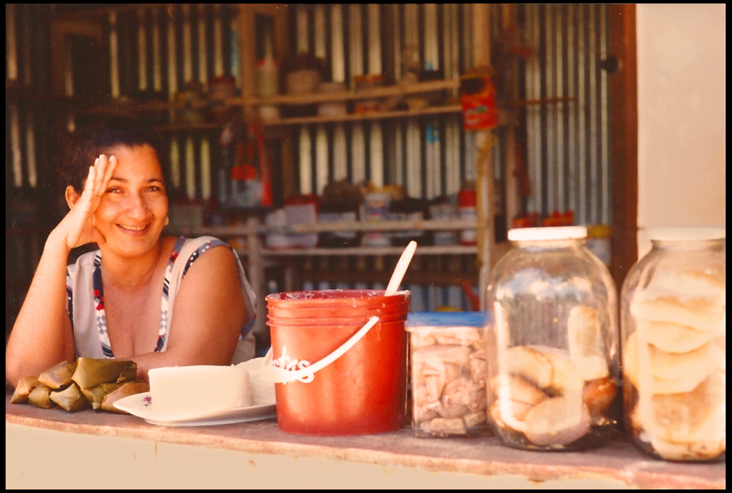 smiling
            middle-aged woman leans on shop counter flanked by home-made
            coste&ntilde;o snack foods in buckets and see-through jars