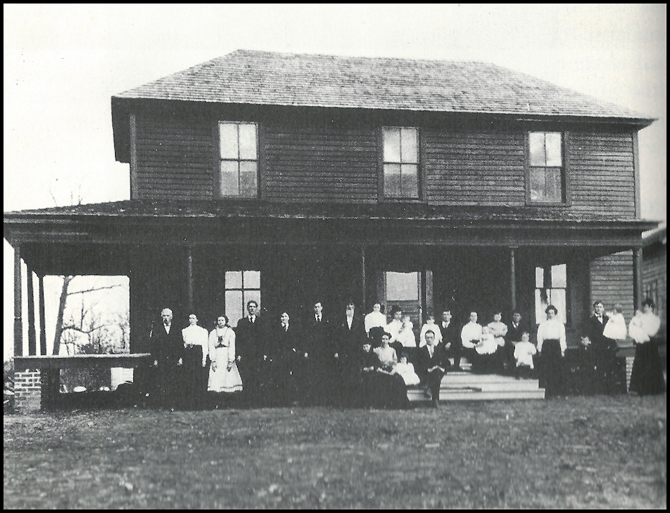 old black and
            white photo: roughly 1900 dark wood frame house with a large
            gathering of people in neat late-Victorian garb