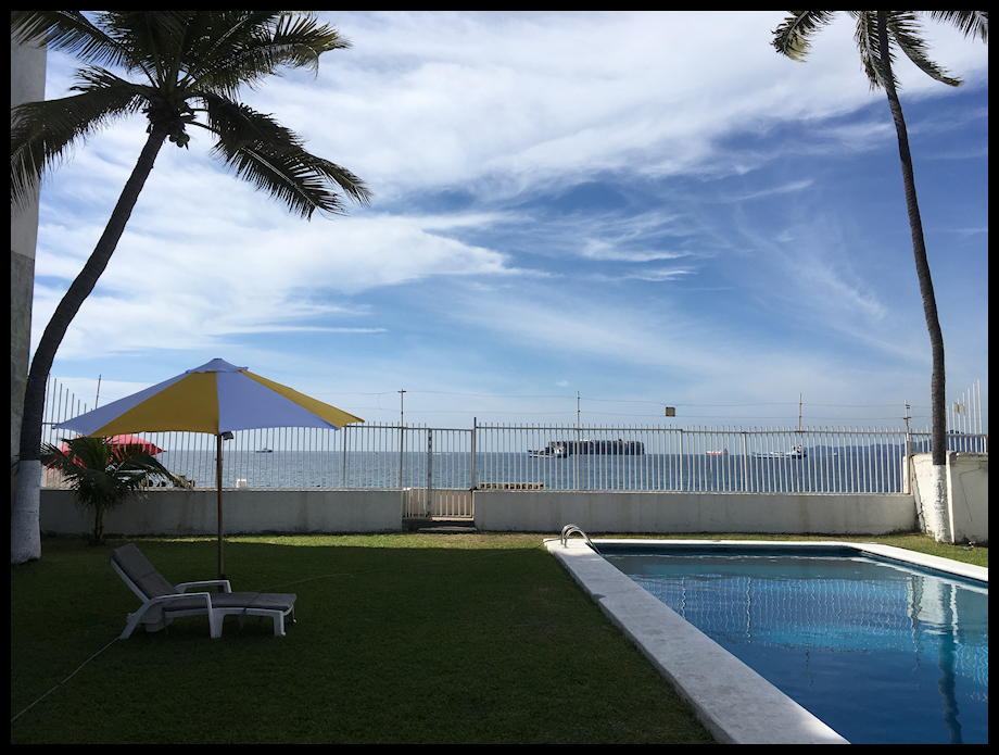 color: pool and lounge chair foreground;
                fence midground; bay with ocean-going vessels, including
                huge laden container ship; Mexican coastal mountains