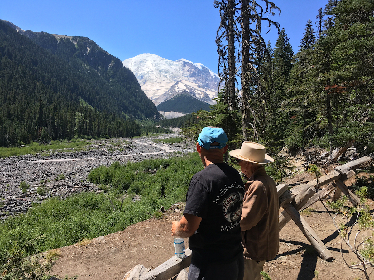 glacier-covered round peak, gorgeously
            clear blue sky, alpine river, fir forest and two men
            admiring the scene