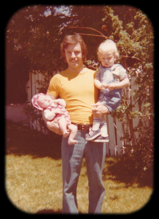 color photo, old and almost
              blurry, 30-ish man standing in yard cradling sleeping
              infant baby girl in right arm while 2-year-old boy sits on
              his left forearm