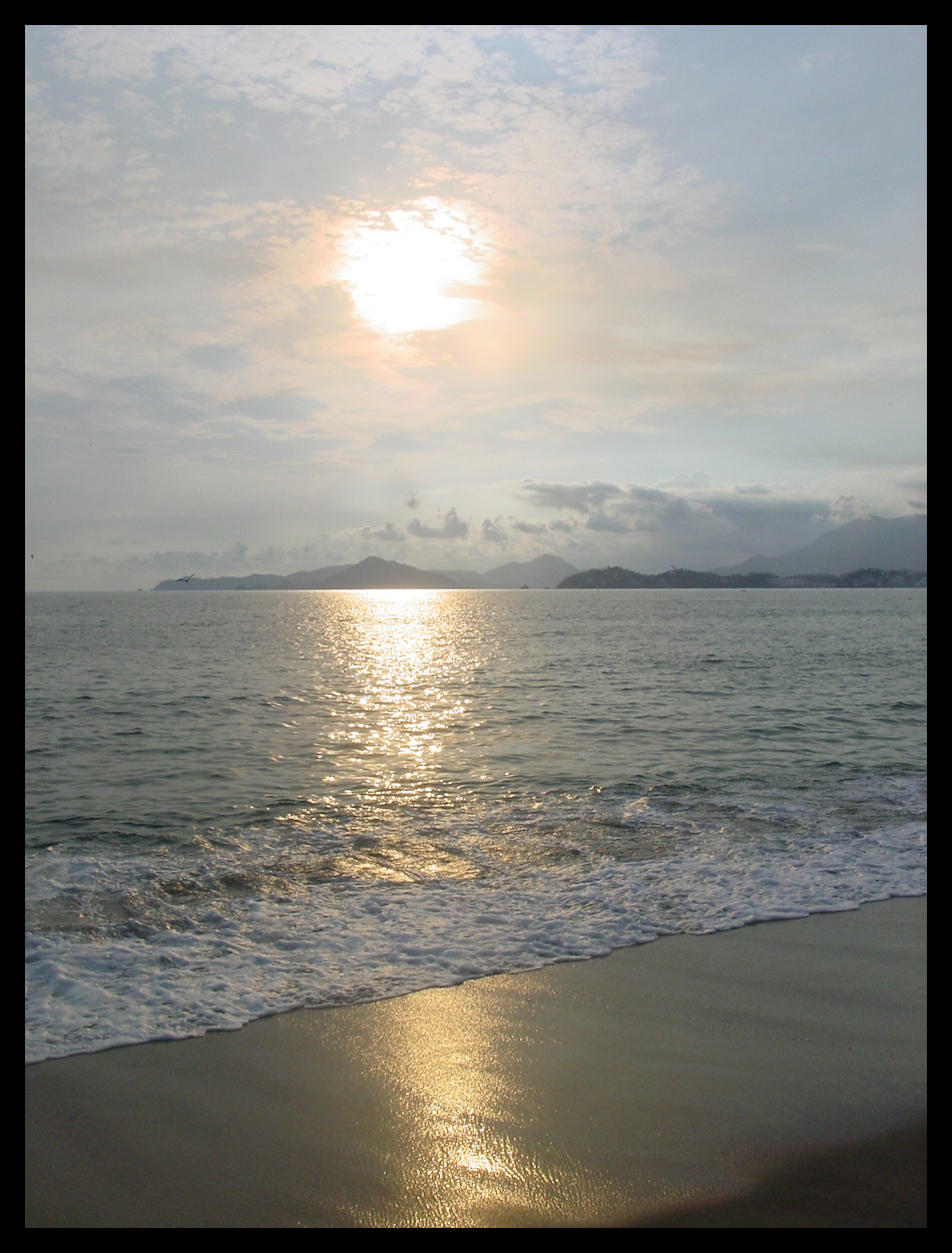 burning hot globe partly
            covered by late-day clouds, hangs over bay, mountains in
            distance, beach and waves in foreground